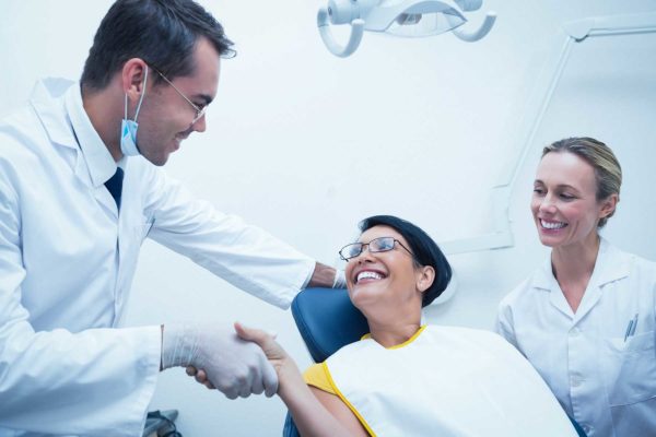 Middle-aged female patient smiling and shaking a male dentist's hand as a female hygienist also smiles at her